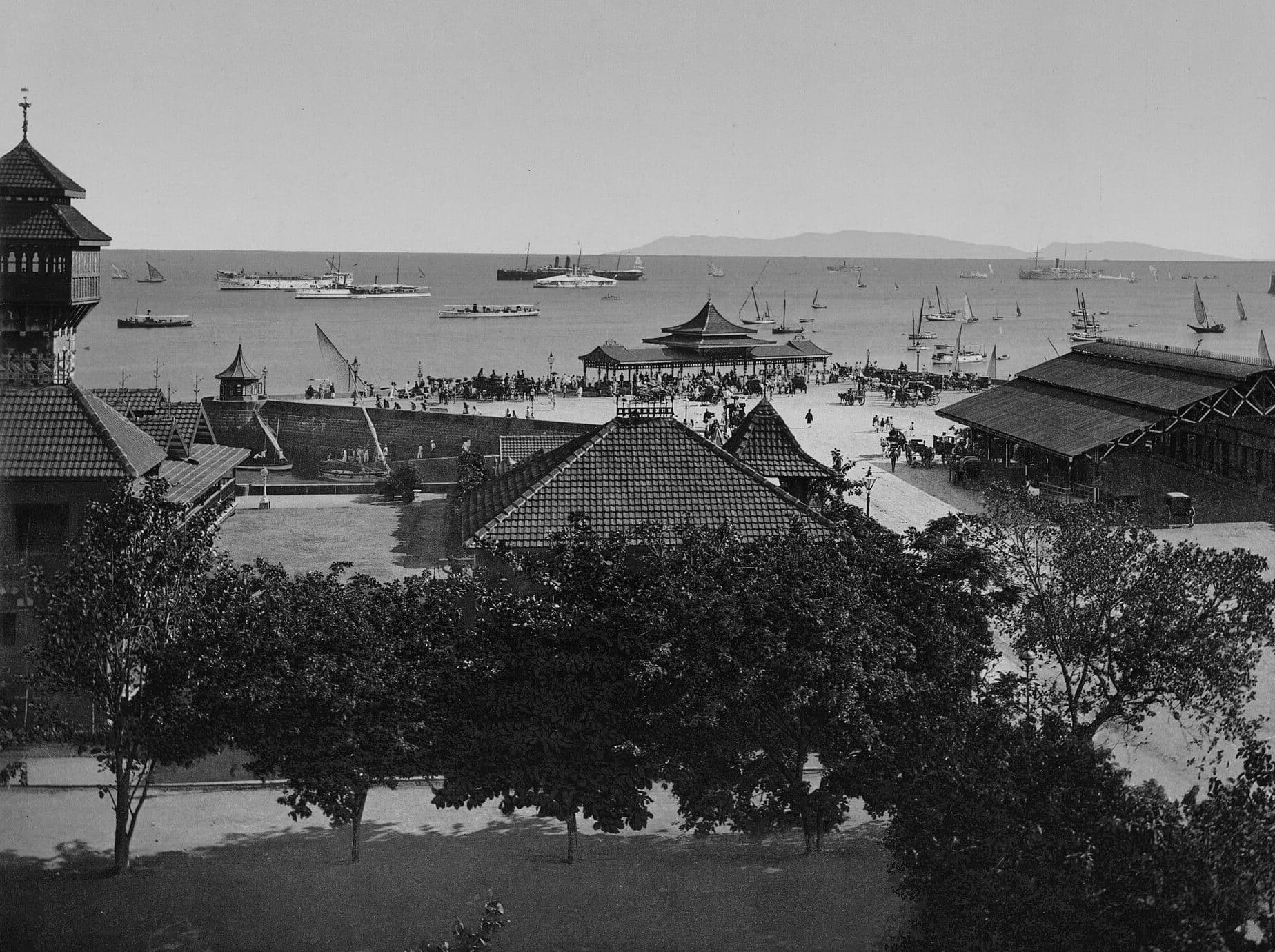 Bombay harbour around 1890: steamships and sailing boats offshore, carriages and crowds on the quay below tiled roofs.